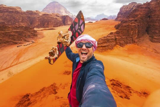 Excited rider pov pose selfie from top viewpoint on big red dune in Wadi Rum desert Jordan. Sand boarding POV with board, desert adventure freedom concept. Wadi rum tours and Sandboarding activities