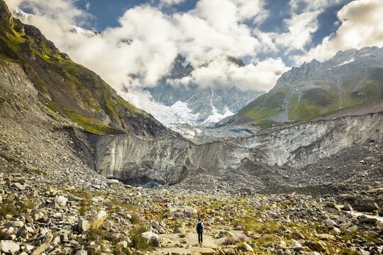 Aerial view woman hiker visit stand by famous hiking destination - Svaneti Shkhara glacier. Summer morning with clouds drift across rugged peaks, moraine rocks foreground. Travel Svaneti explore