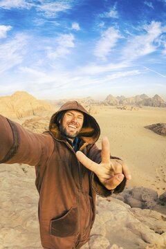 Smiling male tourist traveler wearing hooded traditional brown farwa cloak taking selfie making victory finger gesture vast sand desert valley mountain landscape backdrop Wadi Rum Jordan