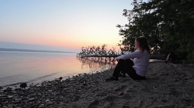 Woman sitting along a peaceful lakeshore at sunrise overlooking a calm lake with soft morning colours in a natural setting.