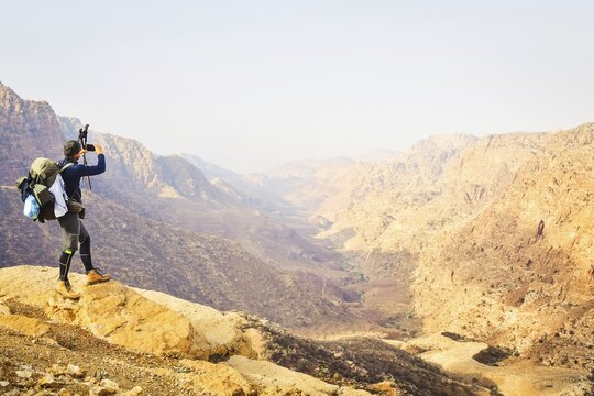 Caucasian Man hiker with large backpack standing rocky ridge taking smartphone picture panoramic sandstone canyon landscape viewpoint hiking travel adventure Dana Reserve Jordan