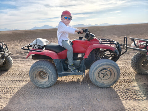 Young adventurer in a red cap confidently rides a red quad bike across the sun drenched desert dunes, embodying the thrill of exploration and the joy of outdoor fun under a bright blue sky.