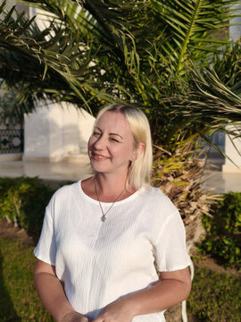 Serene coastal moment a relaxed woman crisp white t shirt smiles gently against backdrop swaying palm trees and tranquil sea, embodying the calm beauty perfect seaside getaway under soft golden sky