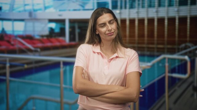 Woman young hispanic standing with crossed arms and pursed lips by indoor swimming pool railing in a public building; skepticism.
