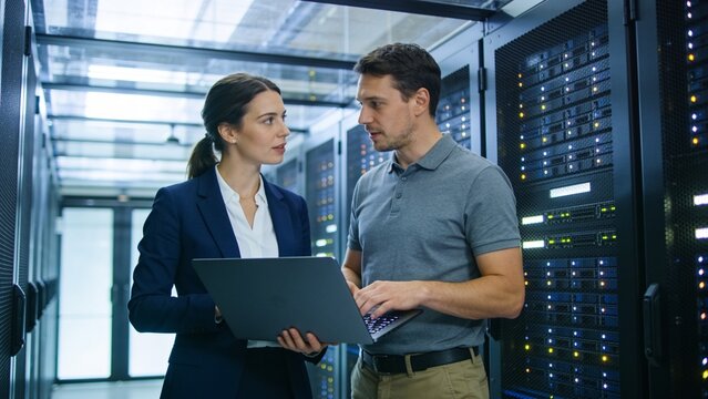 Two cybersecurity engineers collaborating with laptop inside a high-tech cloud computing server room.