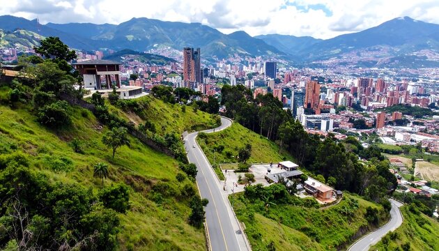 Aerial view of a city nestled between rolling green hills. A winding road leads into the urban expanse, with buildings dotting the landscape