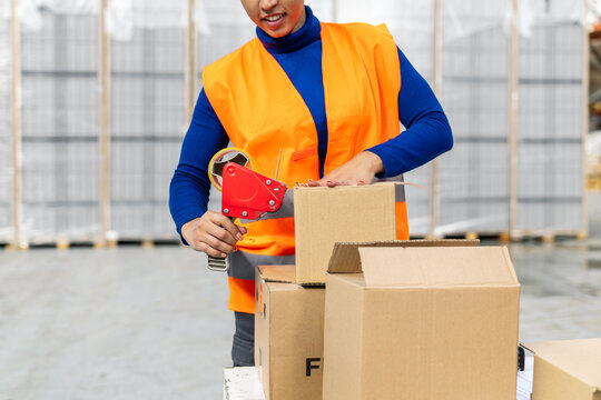 Warehouse worker wearing safety vest packing cardboard boxes using adhesive tape dispenser in a logistics distribution center