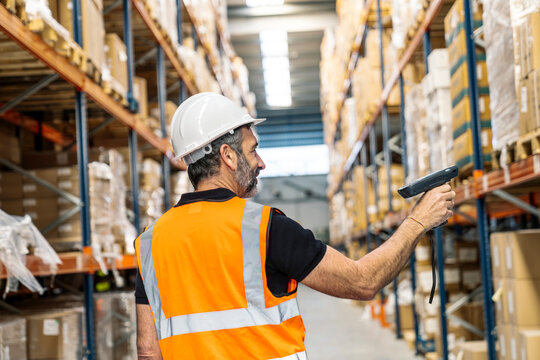 Experienced worker wearing hardhat and safety vest, scanning boxes on shelves in a busy logistics distribution center