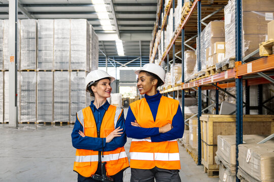 Diverse women logistics workers standing with arms crossed in a large distribution warehouse, smiling and looking at each other