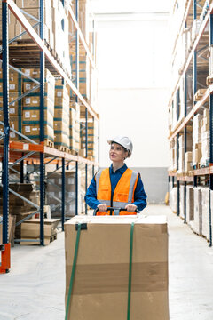 Female worker in safety vest and hard hat pulling cargo box with hand truck inside a modern warehouse logistic center