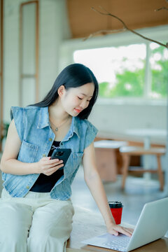 Young Asian woman reading a book with headphones and laptop by the window
