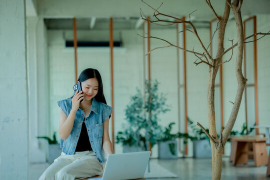 Young Asian woman reading a book with headphones and laptop by the window
