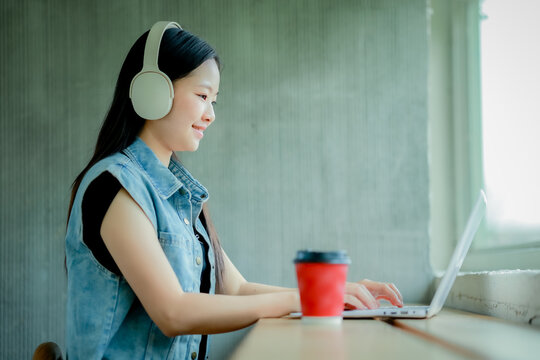 Young Asian woman reading a book with headphones and laptop by the window

