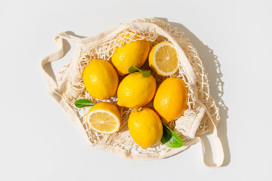 Fresh organic lemons in cotton mesh bag, yellow fruit and green leaves presented on neutral gray background. Organic produce, natural food and healthy shopping themes suit retail and advertising.