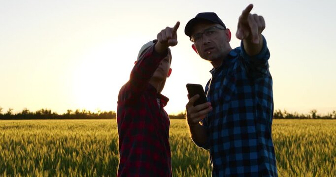 Father and son review information on smartphone in wheat field at sunset, then point toward horizon together. Countryside scene represents guidance, shared vision and preparing future farmer.