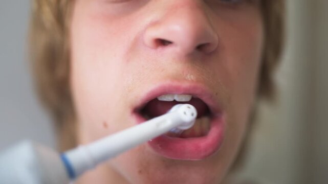 Independent teenage boy brushing his teeth with an electric toothbrush in the bathroom, taking care of his personal hygiene and practicing healthy self-care habits as part of his daily routine.