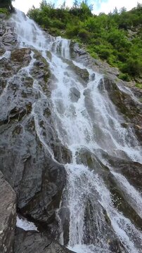 A powerful waterfall cascades down a rocky cliff in the Făgăraș Mountains, Romanian Carpathians, surrounded by lush green forest. Vertical shot ideal for travel and nature cont