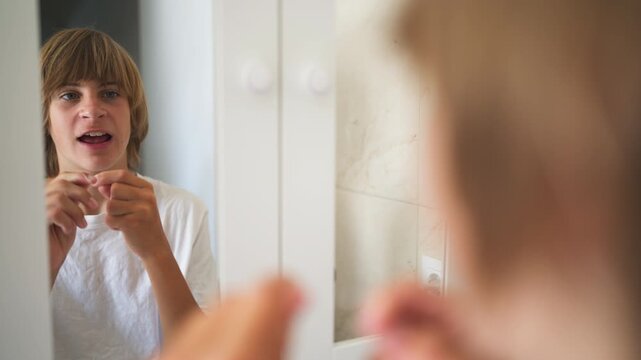 Focused teenage boy flossing his teeth while looking in the bathroom mirror, practicing daily dental hygiene and oral health care as part of his morning personal grooming routine.