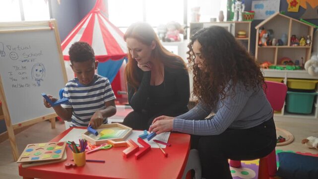 Woman helps boy place wooden puzzle pieces on a red table in a classroom building, guided by a second woman while toys and an easel fill the room; nurturing learning.