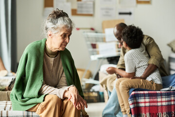 Fototapeta premium Senior Caucasian woman sitting thoughtfully in temporary shelter while Black father comforting child in background, conveying displacement and support. For aid campaigns and social care messaging