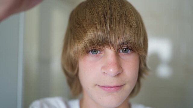 Teenage boy styling fringe with comb while looking into camera and smiling gently, taking care of hairstyle as part of daily grooming and morning routine.