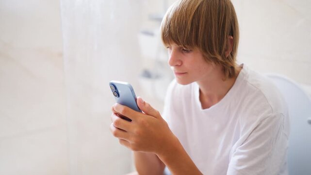 Young boy sitting on the toilet in a bright bathroom while intensely focused on his smartphone, illustrating excessive screen time, digital addiction and smartphone dependency among modern youth.