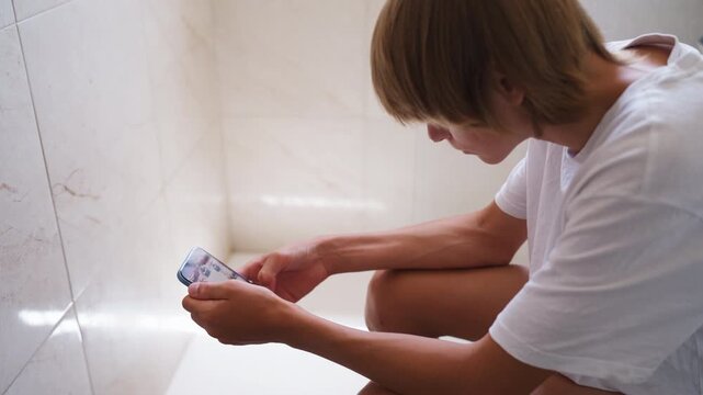 Teenager boy completely absorbed in smartphone while sitting in bathroom, portraying  impact of digital technology, mobile devices and constant connectivity on the daily lives of modern children.