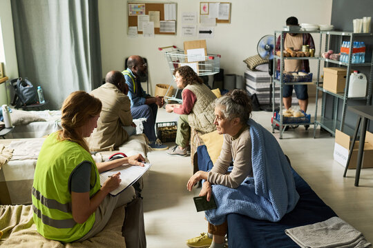 Wide shot of diverse adults receiving shelter support while volunteer registering senior woman in temporary housing center. Ideal for homelessness aid, refugee relief, social care campaigns