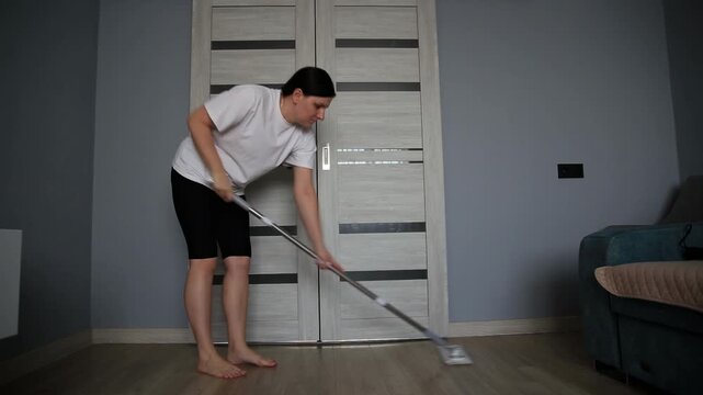 Young adult female cleaning apartment floor with mop, performing household chore, actively engaged in domestic work, maintaining home hygiene, diligent effort in upkeep of living space