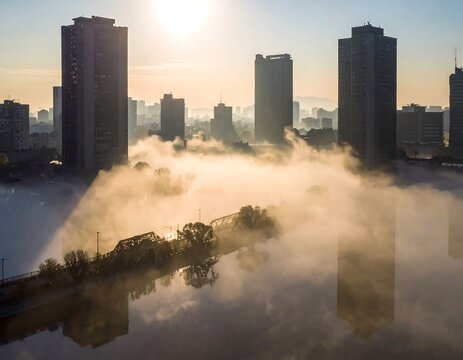 Aerial view of a city skyline shrouded in morning fog, creating a surreal atmosphere with sunlight. Skyscrapers rise above the clouds