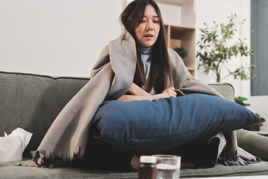 sick asian young woman sneezing into tissue paper while covered with a blanket and sitting on sofa in the living room