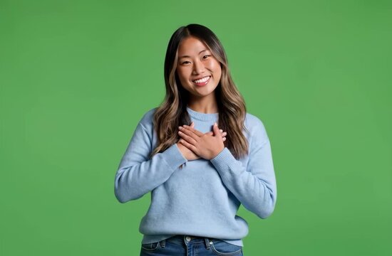 Smiling young Asian woman with hands on chest expressing gratitude and appreciation on a green screen background.