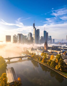 Aerial view of a city skyline shrouded in morning fog, with a river flowing in the foreground under a bright blue sky