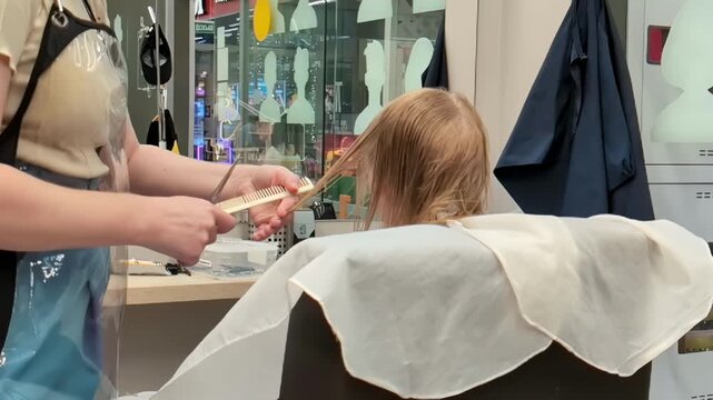 little girl receives hair styling treatment from professional hairstylist in modern hair salon, close up view of hair being cut and combed with scissors and comb, indoors personal care routine