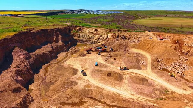Open pit quarry site with photovoltaic panels in background. Wide angle view of rocky landscape, excavators, green fields, solar array, blue sky and clear daylight.