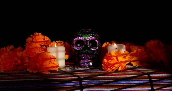 Someone sets down a white candle on a colorful Mexican tablecloth near a painted black metallic skull and red flowers for Dia de los Muertos.