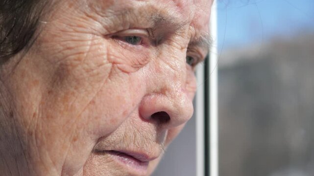 Close up face sad and alone old woman 80 years with deep wrinkled skin looking out window. Gaze filled with nostalgia while reflecting on past life. Woman sick nervous tic and tremor