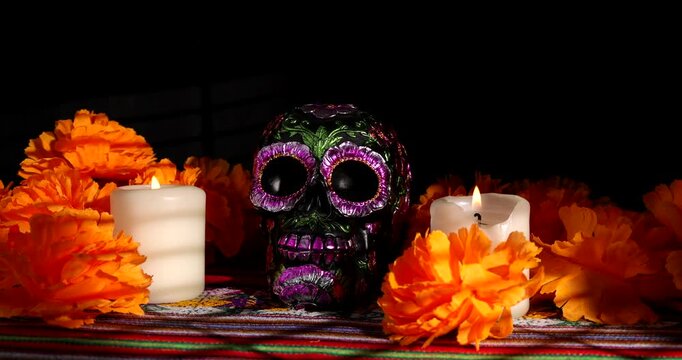 A hand places a white candle near a black metallic painted skull surrounded by red flowers on a traditional Mexican cloth for Day of the Dead.