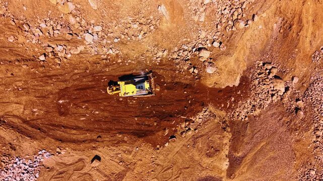 Top down view of bulldozer in stone quarry. Yellow crawler bulldozer moving dark soil and rocks on brown ground surface.