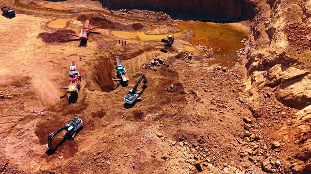 Aerial view of heavy machinery operating in open pit rock quarry. Excavators and crushing machines process extracted stones on reddish sandy soil under bright daylight.