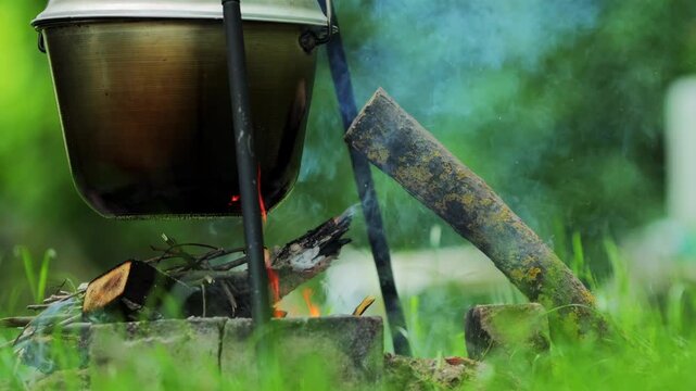 Metal camping pot hangs above burning outdoor campfire, flames and smoke gently rise into air, wood embers glow brightly, natural light illuminates scene slow motion