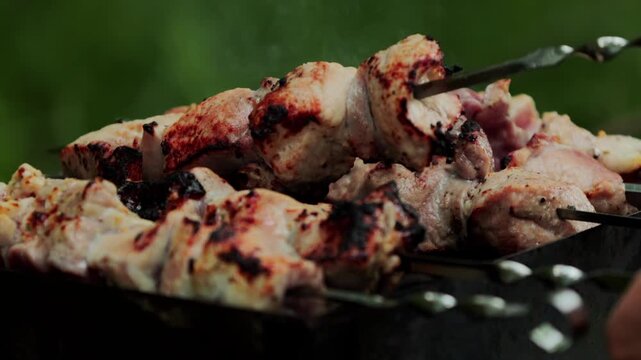 Juicy pork meat pieces cooking on metal skewers over hot charcoal in outdoor brazier grill, smoke rising from delicious traditional shashlik kebab food preparation slow motion