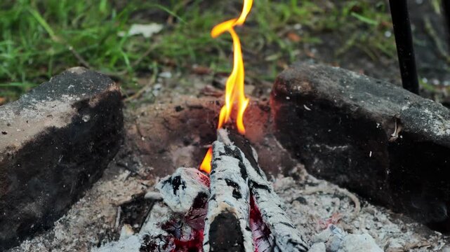 Small campfire burns brightly with flickering orange flames and glowing red embers, wood logs turning to ash, fire pit created with rocks, warmth of summer outdoor recreation slow motion