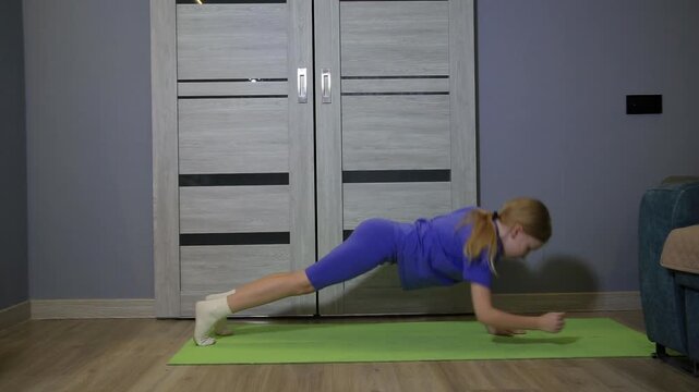 Little girl in blue athletic outfit performing plank exercise on green yoga mat indoors, focused on fitness and strength training for healthy lifestyle at home