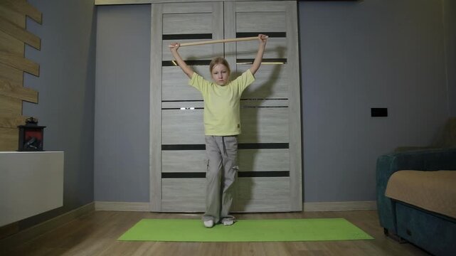 Young girl performs physical exercise with wooden stick held above head, standing on yoga mat indoors, focused on fitness routine and healthy lifestyle, at home during day