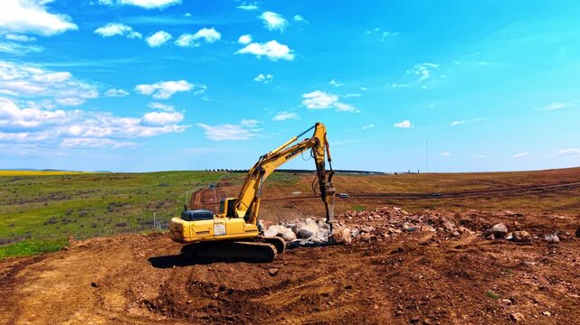 Yellow crawler excavator breaking rocks with hydraulic hammer at quarry. Heavy machine with jackhammer works on brown rocky soil under blue sky.