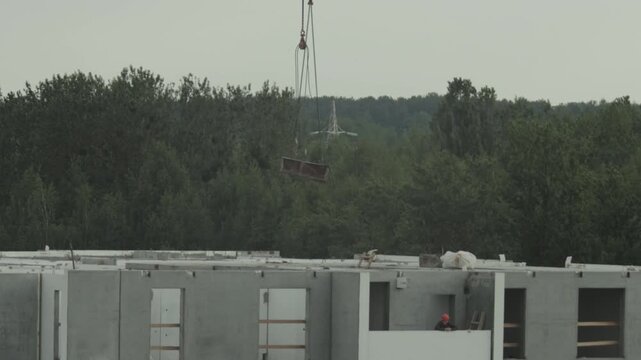Construction site tower crane moving building material above concrete structure, worker in hard hat observing progress residential development