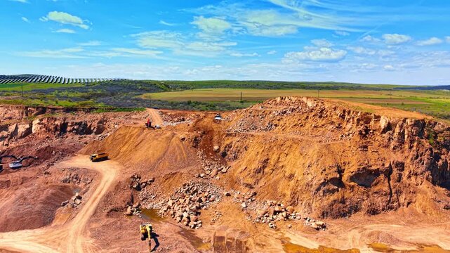 Wide open pit rock quarry with solar farm on horizon. Red earth mining area with trucks and excavators under clear blue sky with green hills.