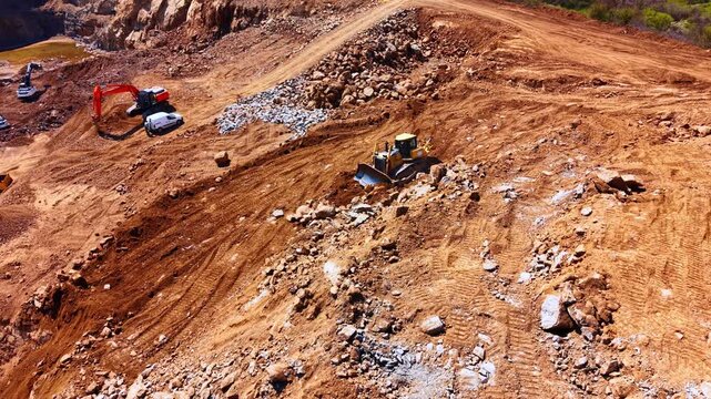 Aerial view of heavy equipment operating in open pit quarry. Excavator and bulldozer work among stone piles on red terraced terrain under sunlight.
