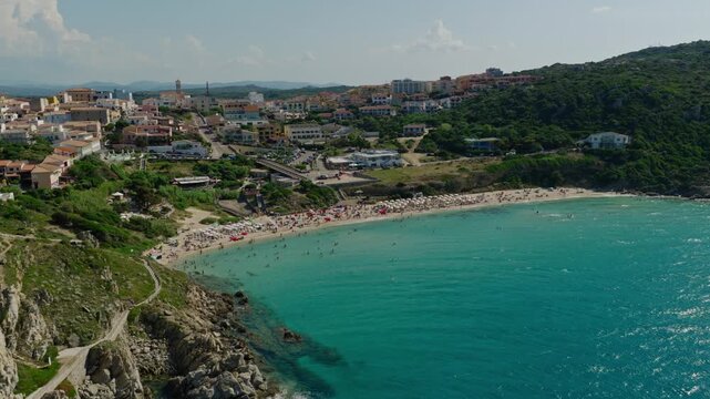 Drone flight above Rena Bianca Beach in Santa Teresa Gallura, revealing turquoise Mediterranean water, a lively sandy shore, rocky cliffs and the coastal town.